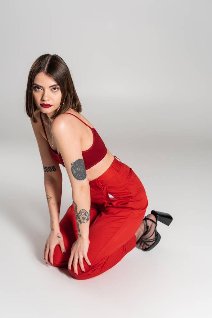 mladý model v červeném oblečení, tetovaná brunetka žena s krátkými vlasy a nosem piercing pózování v červené plodině top a kalhoty na šedém pozadí, moderní styl, generace z, módní trend  - Fotografie, Obrázek