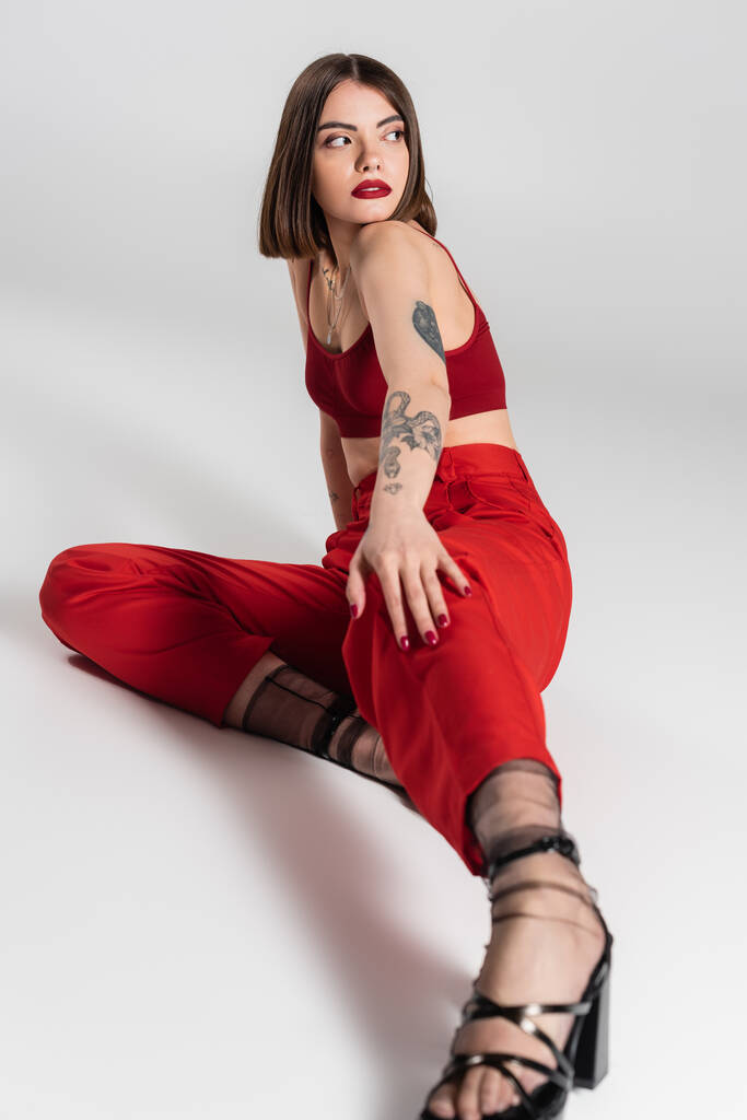 divatos ruha, fiatal modell piros ruhában, magabiztos és tetovált nő rövid haj és orr piercing pózol piros növény felső és nadrág szürke háttér, generáció z, teljes hossz  - Fotó, kép