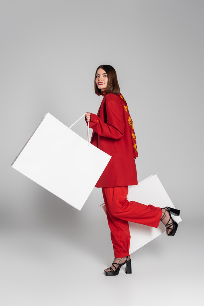 потребительство, счастливая женщина с брюнеткой короткие волосы и нос пирсинг держа сумки и ходить на сером фоне, современный модный тренд, модный наряд, красный костюм, полная длина  - Фото, изображение