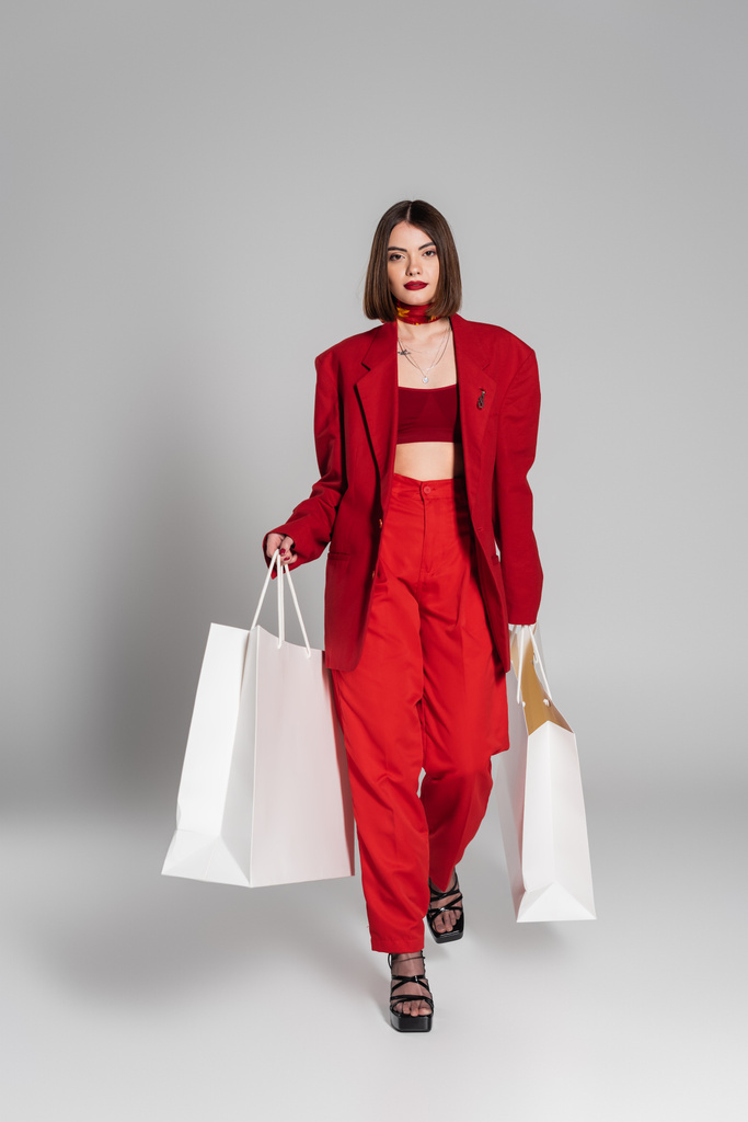 consumerism, young woman with brunette short hair and nose piercing holding shopping bags and walking on grey background, modern fashion trend, fashionable outfit, youth culture, full length  - Photo, Image