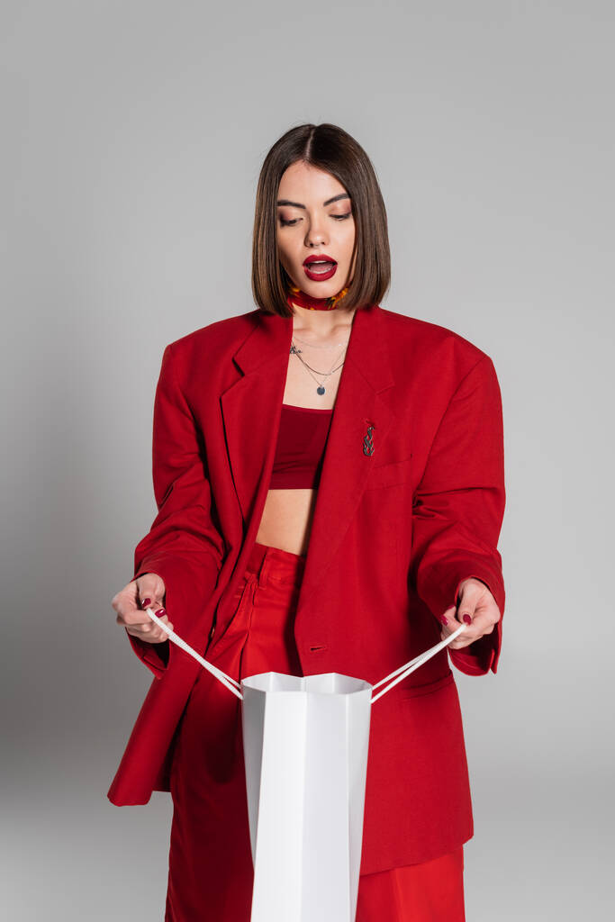 fashionable, generation z, shocked young woman with brunette short hair and nose piercing looking inside of shopping bag on grey background, youth culture, red suit, consumerism  - Photo, Image