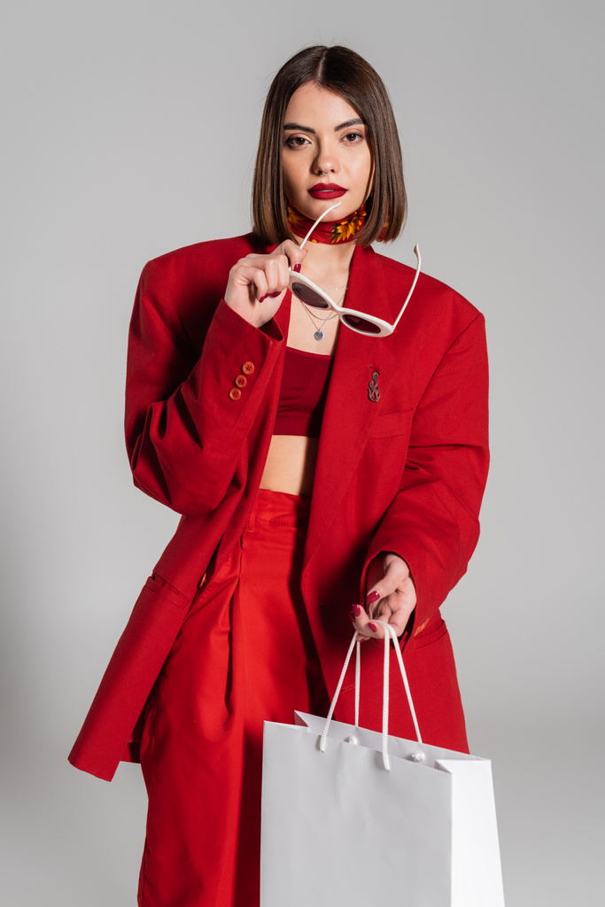 μοντέρνα, γενιά z, σοκαρισμένη νεαρή γυναίκα με κοντά καστανά μαλλιά και σκουλαρίκια στη μύτη κρατώντας γυαλιά ηλίου και τσάντα για ψώνια σε γκρι φόντο, κουλτούρα της νεολαίας, κόκκινο κοστούμι, καταναλωτισμός  - Φωτογραφία, εικόνα