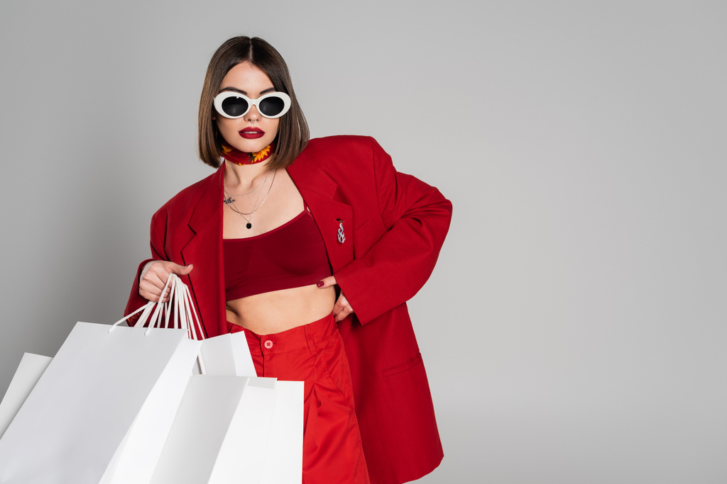 fashionable, generation z, young woman with brunette short hair and nose piercing posing in sunglasses and red suit while holding shopping bags on grey background, youth culture, consumerism  - Photo, Image