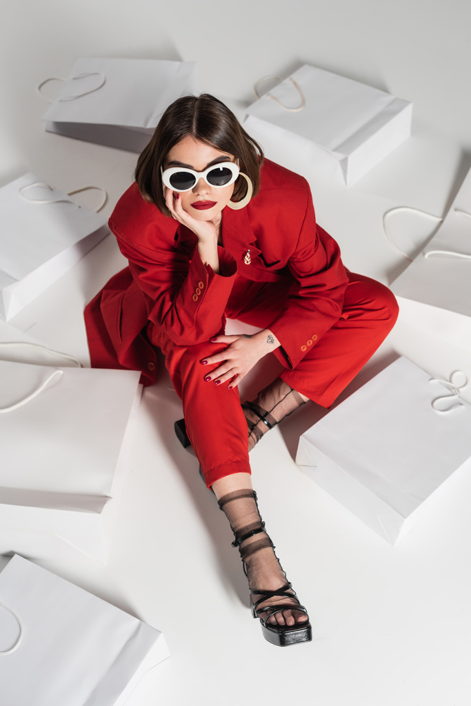 consumismo, mujer joven con pelo corto morena, piercing en la nariz y tatuaje posando en gafas de sol y traje rojo mientras se sienta alrededor de bolsas de compras sobre fondo gris, vista de ángulo alto  - Foto, Imagen
