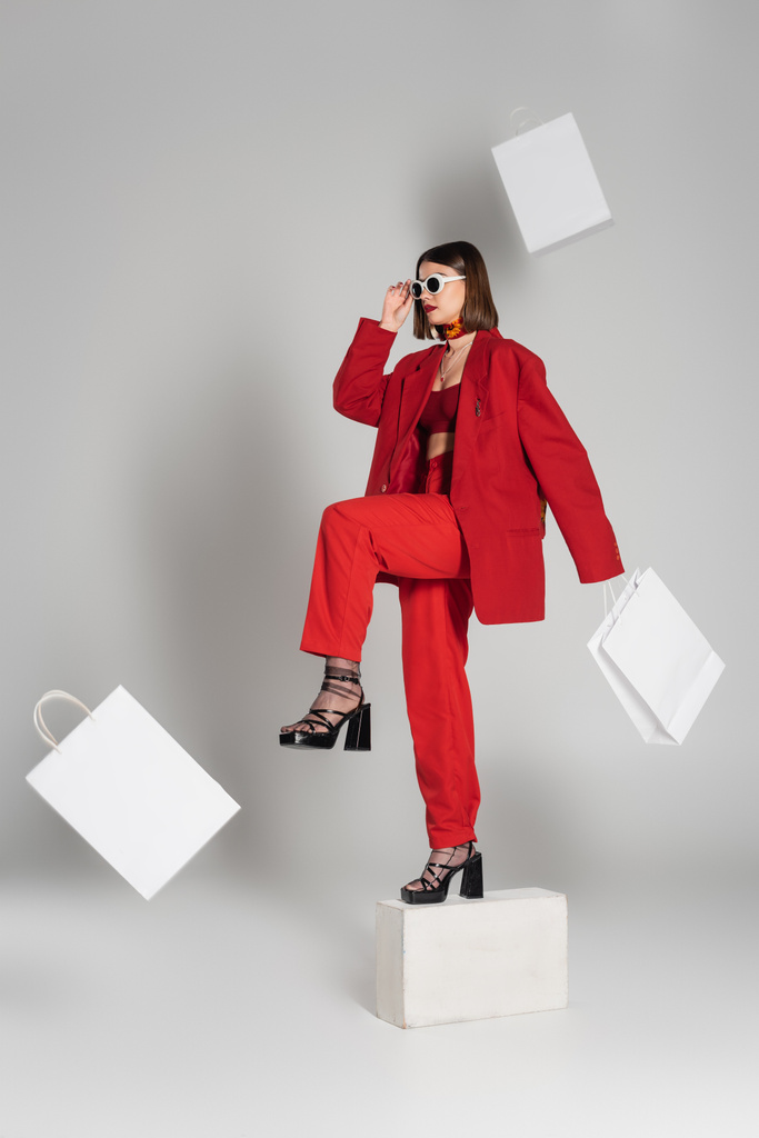 Generation Z, Konsum, Modemodel mit brünetten kurzen Haaren und Nasenpiercing posiert in Sonnenbrille und rotem Anzug auf Betonwürfeln um fliegende Einkaufstüten auf grauem Hintergrund  - Foto, Bild