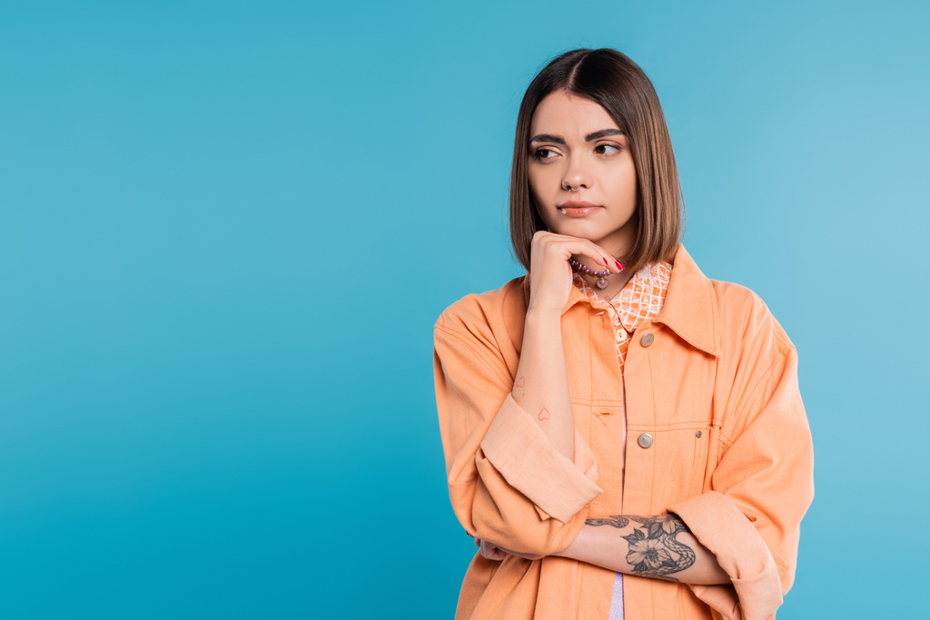 generation z, portrait of concerned woman, young fashion model looking away and thinking on blue background, orange shirt, short brunette hair, pierced nose, summer outfit, gen z fashion  - Photo, Image