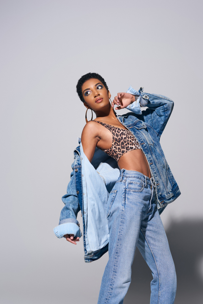 Fashionable african american model with short hair, vibrant makeup and golden earrings wearing denim jacket, top and jeans while standing on grey background, denim fashion concept - Photo, Image