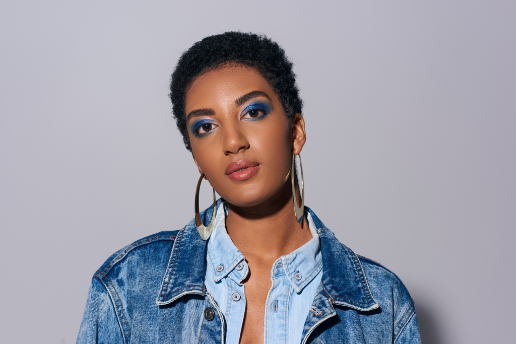 Portrait of modern and confident young african american woman with bold makeup and golden earrings wearing denim outfit and looking at camera on grey background, denim fashion concept - Photo, Image