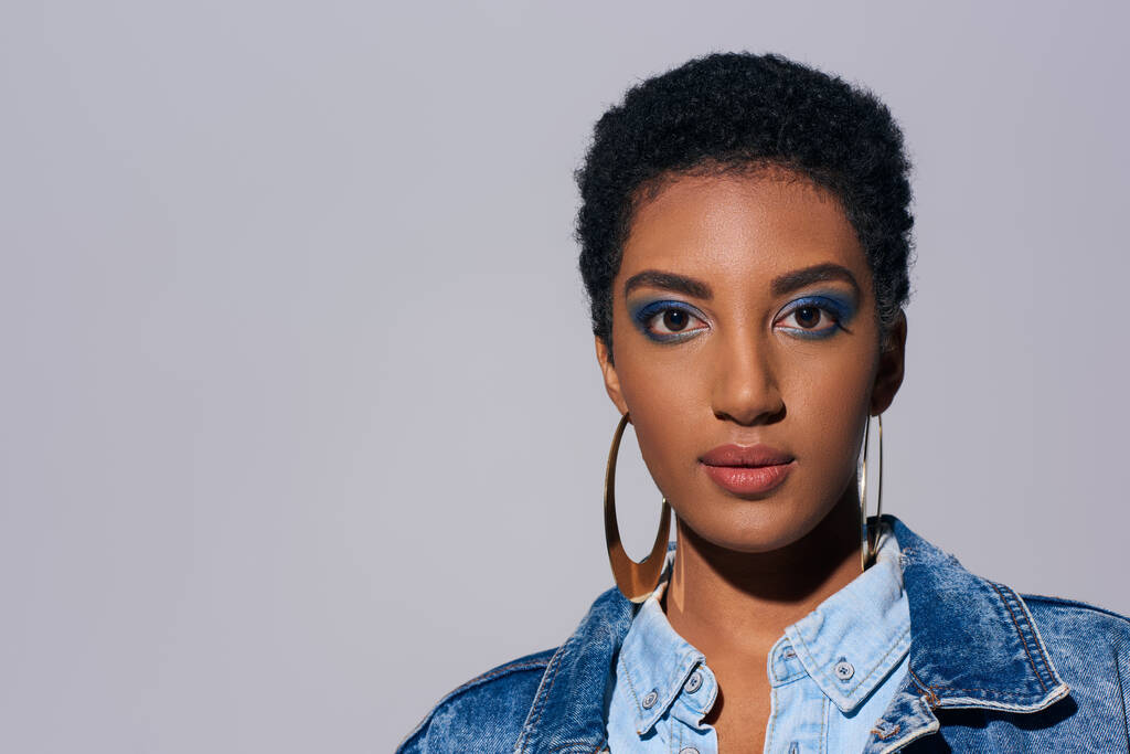 Confident young african american woman with vivid makeup and golden earrings wearing denim outfit and looking at camera isolated on grey, denim fashion concept - Photo, Image