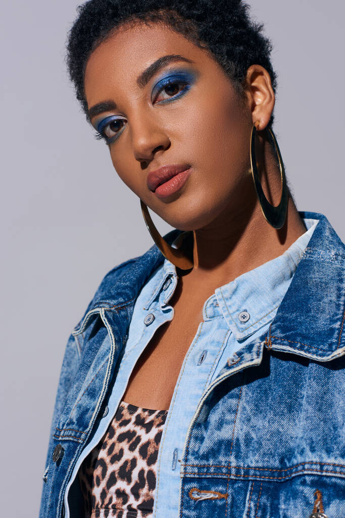 Mulher americana africana de cabelos curtos e confiante com maquiagem ousada e brincos dourados vestindo top e traje de ganga enquanto está isolada no conceito de moda cinza e jeans - Foto, Imagem
