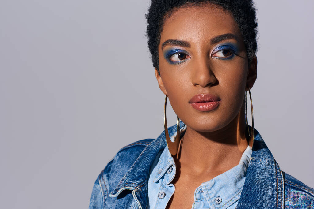 Retrato da jovem afro-americana de cabelos curtos e elegantes com maquiagem vívida e jaqueta de ganga olhando para longe enquanto estava isolada no conceito de moda cinza e jeans - Foto, Imagem