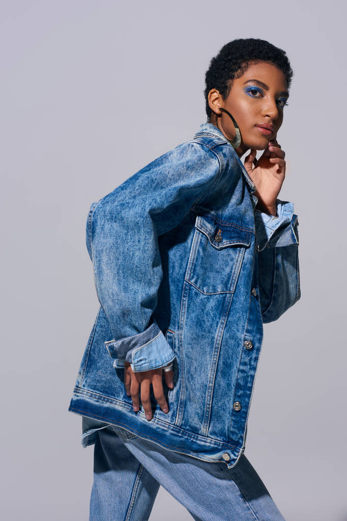 Modernes afrikanisch-amerikanisches Model mit fettem Make-up und kurzen Haaren, die das Kinn berühren und in die Kamera schauen, während sie im Jeans-Outfit isoliert auf grauem, Denim-Modekonzept posieren - Foto, Bild