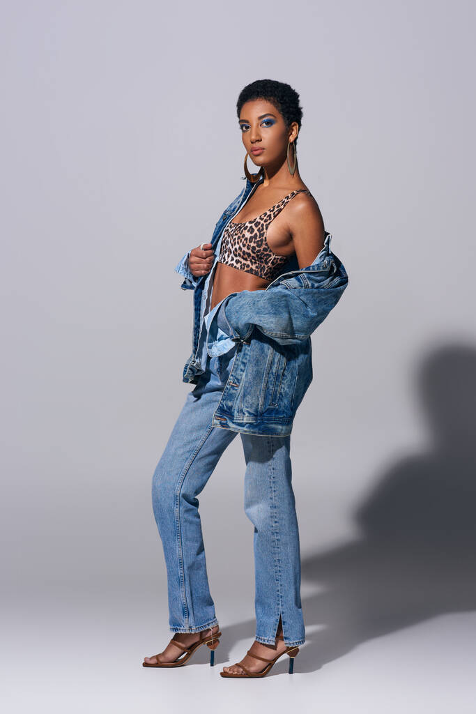 Volle Länge der modischen afrikanisch-amerikanischen Frau mit fettem Make-up trägt Top mit Animal Print, Jeansjacke und Jeans auf grauem Hintergrund, Denim-Modekonzept - Foto, Bild