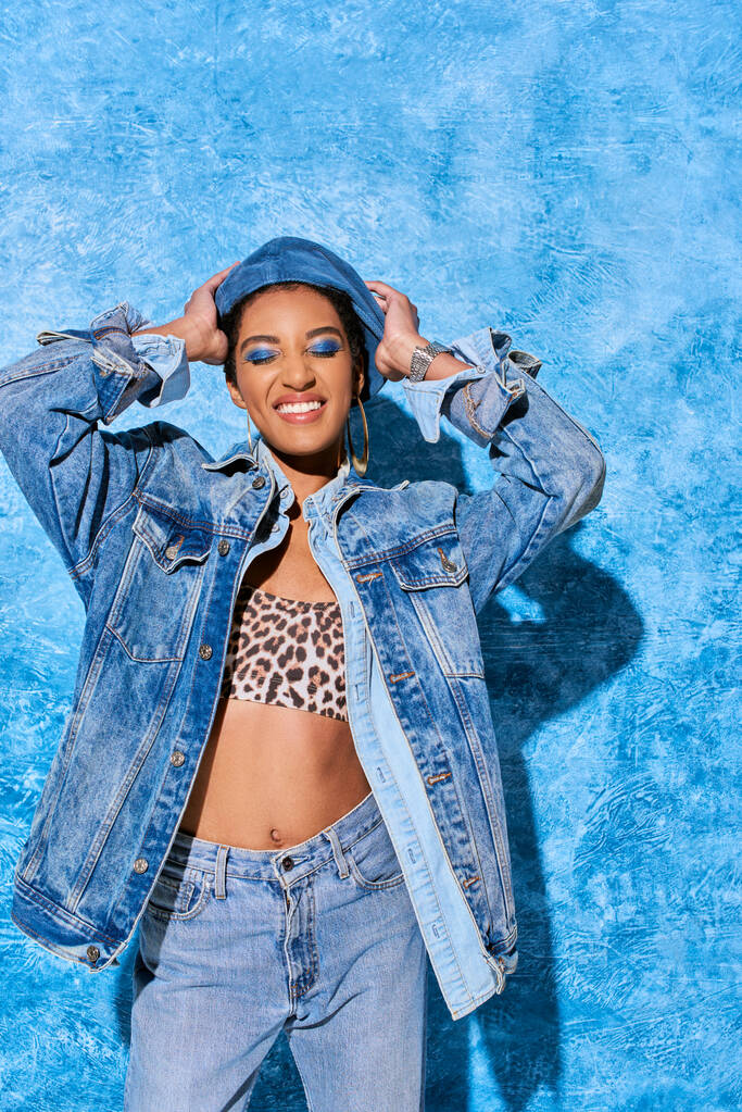 Femme afro-américaine à la mode avec les yeux fermés et le maquillage audacieux touchant béret et posant dans des vêtements en denim et haut sur fond texturé bleu, tenue en denim élégant - Photo, image