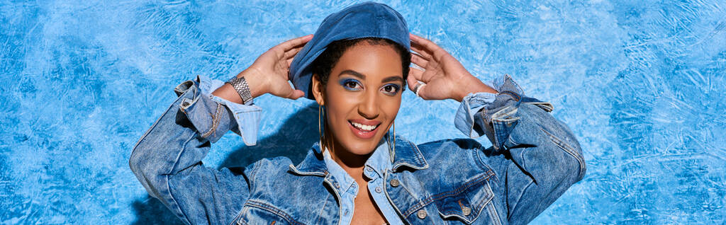 Porträt eines lächelnden und kurzhaarigen afrikanisch-amerikanischen Models, das Baskenmütze berührt, während es in Jeansjacke posiert und auf blauem strukturiertem Hintergrund steht, stylische Jeans-Kleidung, Banner  - Foto, Bild