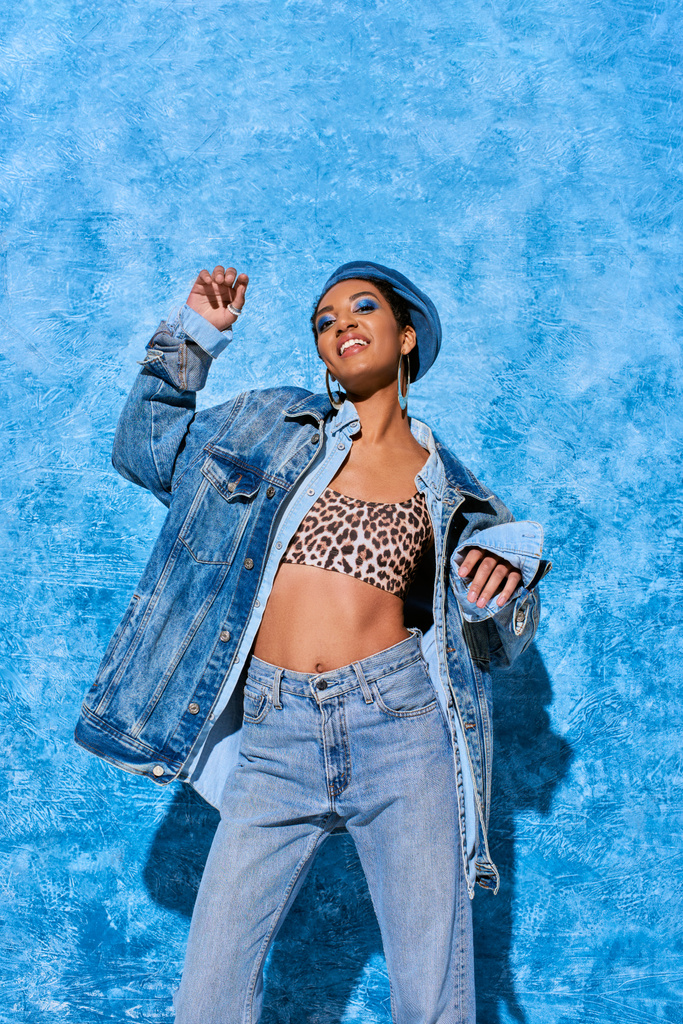Baixo ângulo de visão da moda jovem modelo afro-americano na boina posando em cima e roupas de ganga enquanto sorri para a câmera em fundo azul texturizado, traje de ganga elegante - Foto, Imagem
