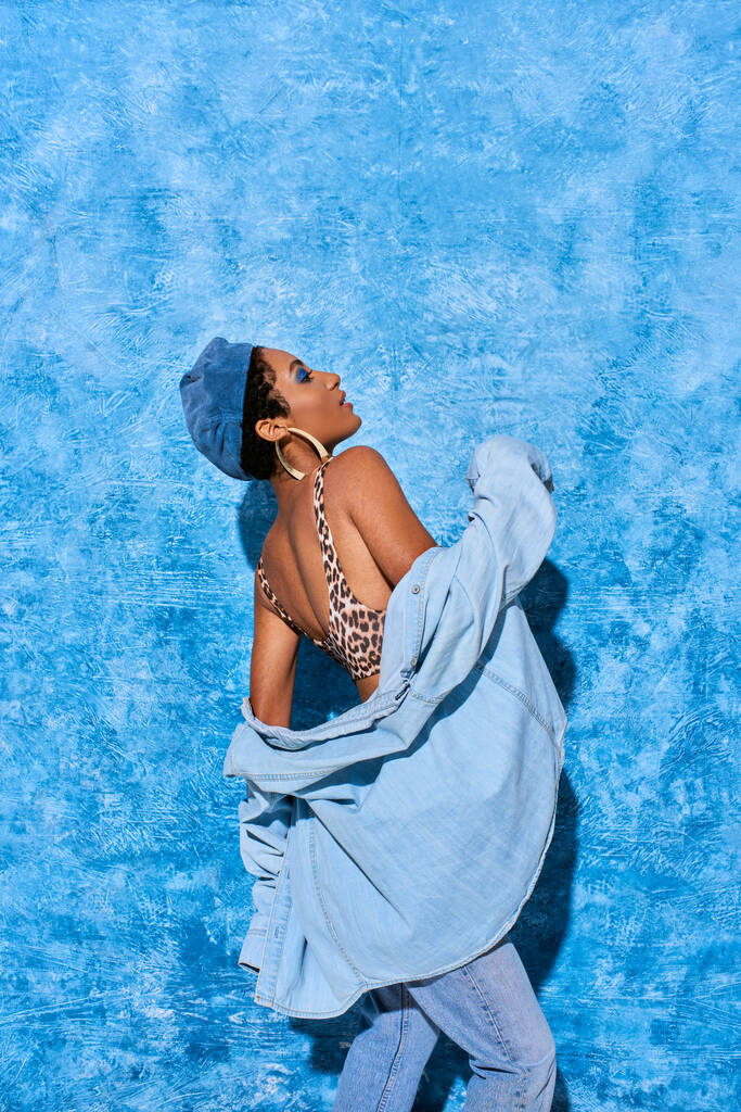 Zijaanzicht van modieuze Afrikaanse Amerikaanse vrouw in baret, top met dierenprint en denim shirt poseren en staan op blauwe textuur achtergrond, stijlvolle denim kleding - Foto, afbeelding