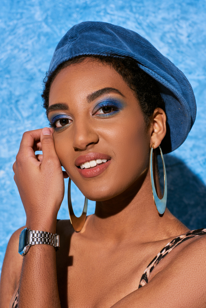 portret van jonge Afrikaans-Amerikaanse vrouw met gedurfde make-up, baret en gouden oorbellen glimlachen op de camera en poseren op blauwe textuur achtergrond, stijlvolle denim kleding - Foto, afbeelding