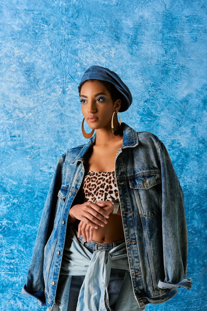 Trendy african american model in beret and golden earrings posing in denim jacket and top with animal pattern while standing on blue textured background, stylish denim attire - Photo, Image