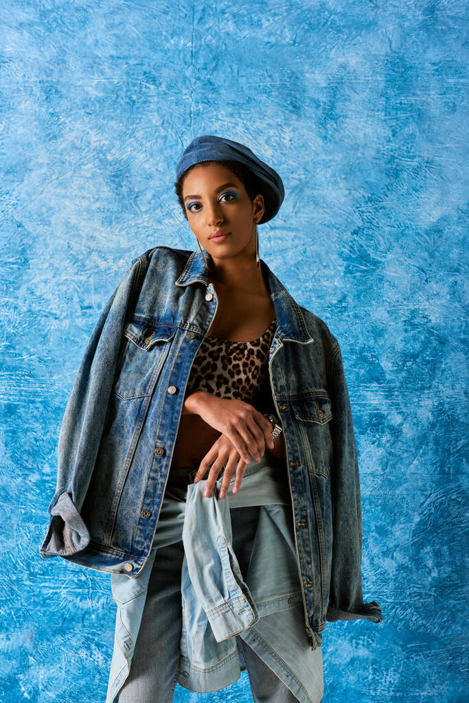 Молодая африканская американка в берете смотрит в камеру, позируя в джинсовой одежде и топе с рисунком животного на синем текстурированном фоне, стильный джинсовый наряд - Фото, изображение