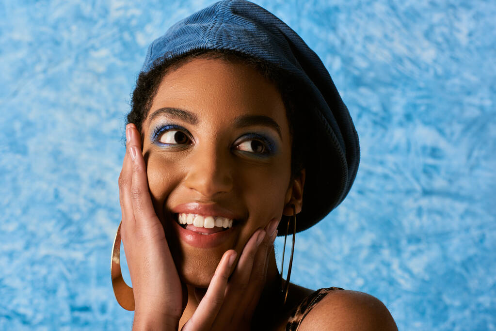 Portret van positief Afrikaans Amerikaans model met levendige make-up, baret en oorbellen aanraken wangen en kijken weg op blauwe textuur achtergrond, stijlvolle denim kleding - Foto, afbeelding