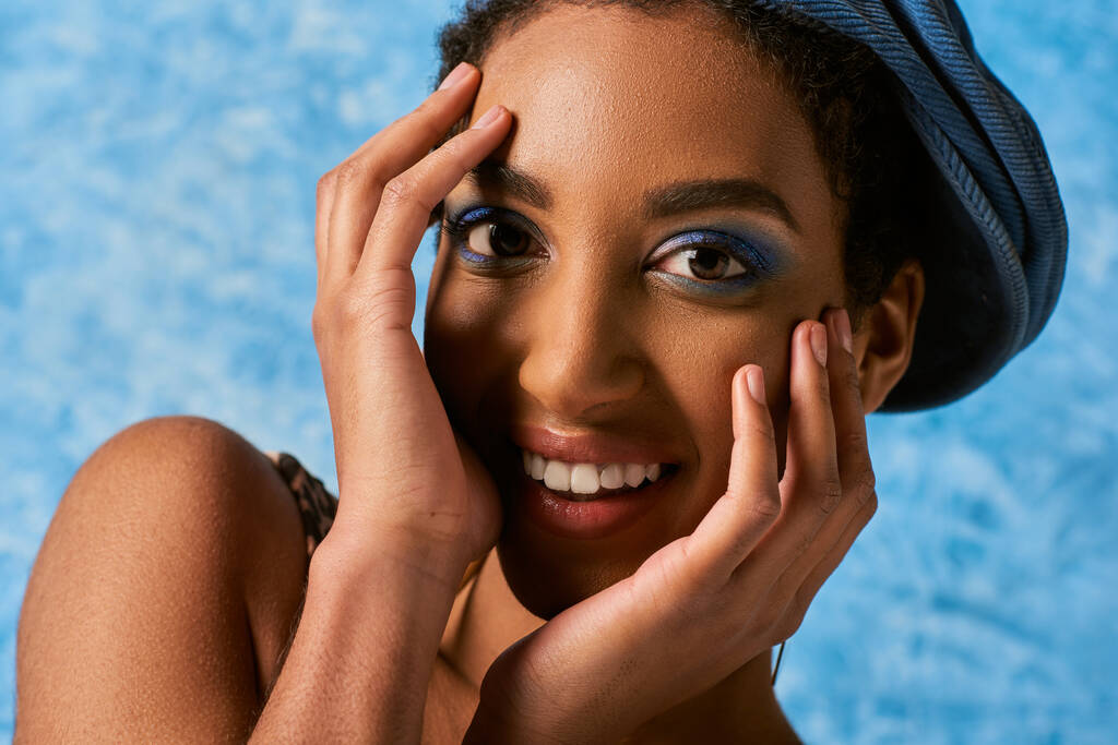 Close-up vista de sorrir e jovem mulher afro-americana com maquiagem vívida e boina tocando rosto e olhando para a câmera em fundo azul texturizado, traje de ganga elegante - Foto, Imagem