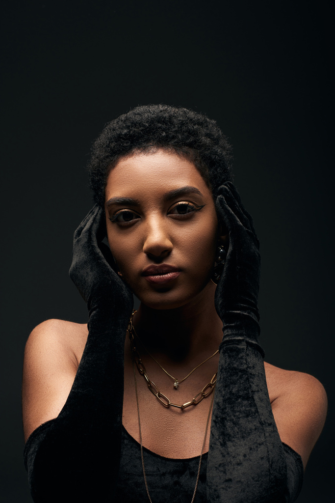 Portrait of fashionable and short haired african american woman with makeup and golden necklaces wearing gloves and dress while standing isolated on black, high fashion and evening look - Photo, Image