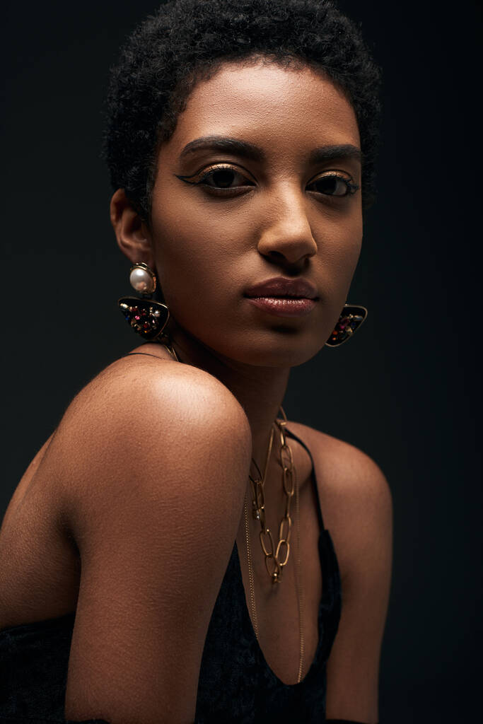 Elégant jeune modèle afro-américain en robe de soirée, maquillage et accessoires dorés regardant la caméra isolée sur noir avec éclairage, look haute couture et soirée - Photo, image
