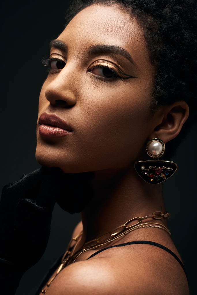 Vista de cerca de la joven mujer afroamericana segura y elegante con maquillaje de noche y accesorios dorados mirando a la cámara aislada en negro, alta moda y aspecto de noche - Foto, imagen