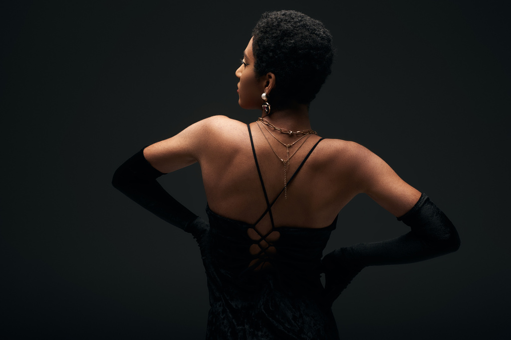 Oldalnézet divatos fiatal afro-amerikai nő elegáns kesztyű és arany kiegészítők fogják egymás kezét a csípő elszigetelt fekete, magas divat és esti megjelenés - Fotó, kép