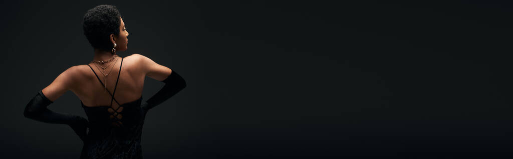 Vista lateral del modelo afroamericano de moda en guantes elegantes y accesorios dorados que tocan las caderas mientras están de pie aislados en negro, alta moda y aspecto de noche, pancarta  - Foto, imagen