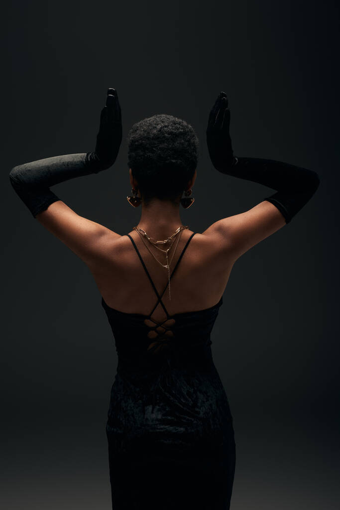 Rückansicht einer eleganten afrikanisch-amerikanischen Frau in Abendkleid, Handschuhen und goldenen Accessoires posiert isoliert auf schwarz mit Beleuchtung, High Fashion und Abendlook, feminine Sinnlichkeit  - Foto, Bild
