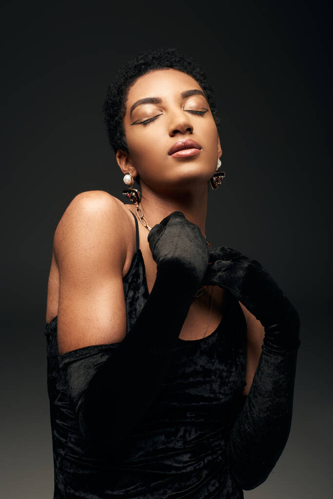 Portrait de femme afro-américaine aux cheveux courts à la mode et élégante avec maquillage, accessoires dorés et gants debout les yeux fermés isolés sur look noir, haute couture et soirée - Photo, image