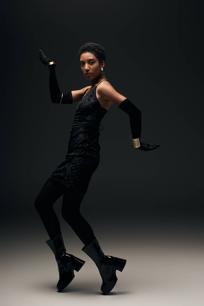 Full length of confident young african american model in little dress, γάντια και μπότες ποζάρουν σε μαύρο φόντο με φωτισμό, υψηλή μόδα και βραδινό look, μοντέλο μόδας  - Φωτογραφία, εικόνα