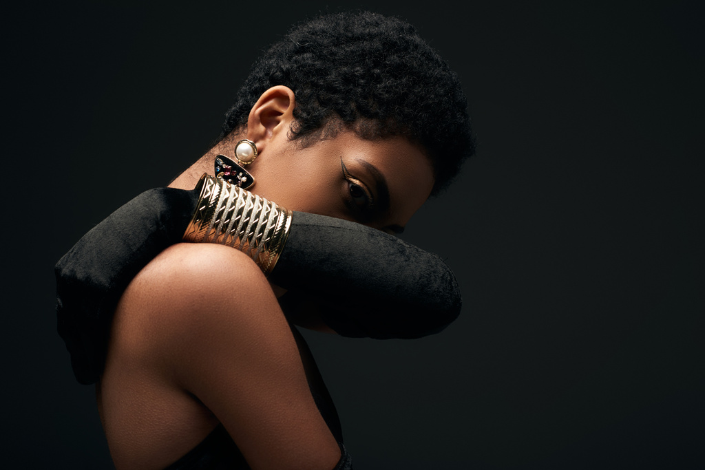 Fashionable african american woman with evening makeup wearing accessories and glove while covering face and looking at camera isolated on black, high fashion and evening look - Photo, Image