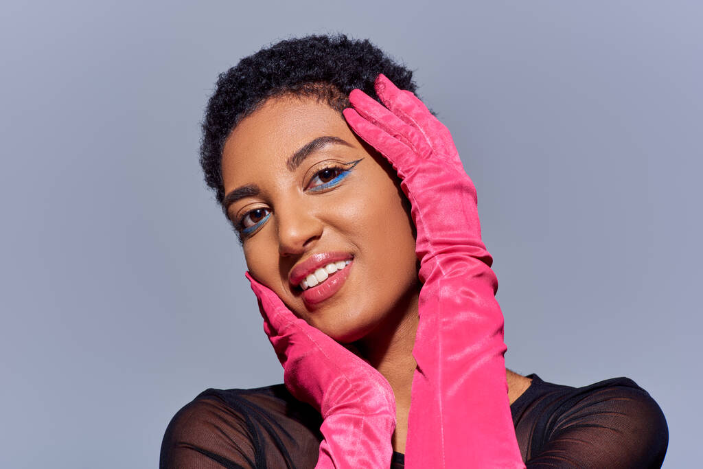 Muotokuva positiivinen ja lyhytkarvainen afrikkalainen amerikkalainen nainen rohkea meikki ja vaaleanpunainen käsineet koskettaa kasvot ja katsot kameran eristetty harmaa, moderni sukupolvi z muoti käsite - Valokuva, kuva