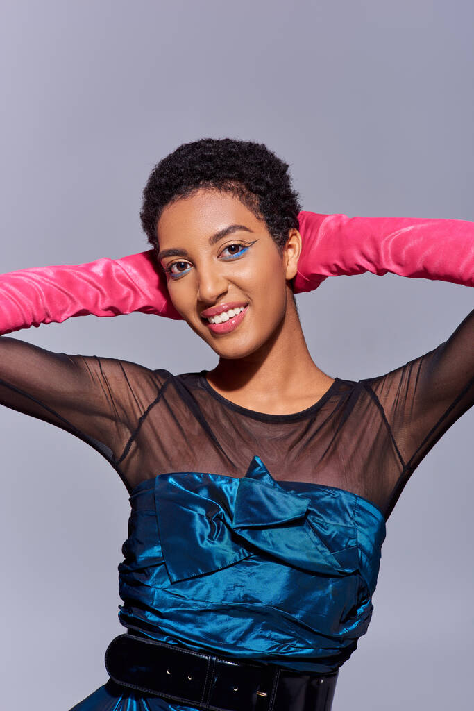 Smiling and fashionable african american woman with bold makeup wearing pink gloves and cocktail dress and touching head isolated on grey, modern generation z fashion concept - Photo, Image