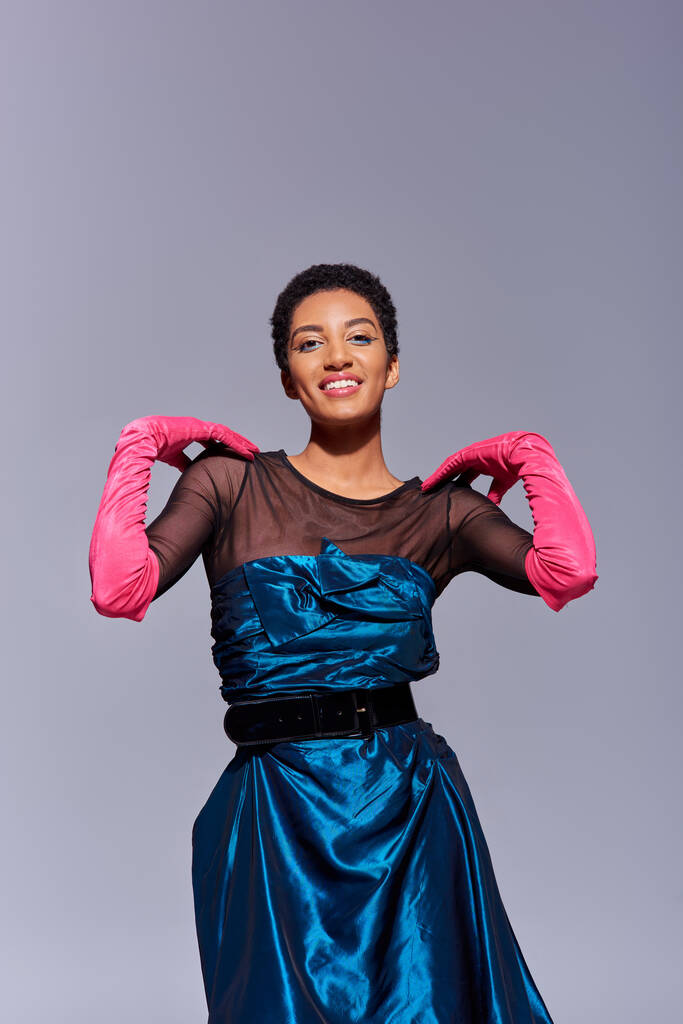 Positive african american woman in cocktail dress and pink gloves touching shoulders and looking at camera isolated on grey, modern generation z fashion concept - Photo, Image