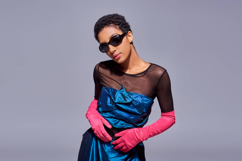 Sebevědomý mladý a elegantní africký americký model v slunečních brýlích, koktejlových šatech a růžových rukavicích pózujících izolovaně na šedé, moderní generace z módní koncepce - Fotografie, Obrázek