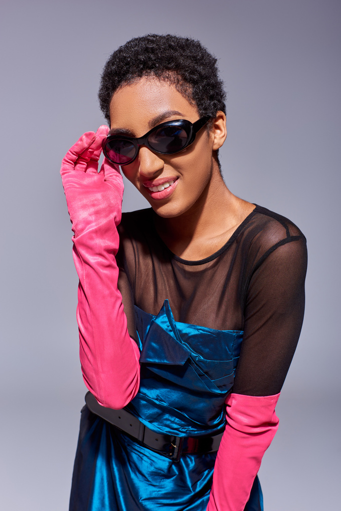 Retrato de jovem sorridente e de cabelos curtos modelo americano africano em luvas rosa e vestido de cocktail tocando óculos de sol isolados em cinza, conceito de moda z geração moderna - Foto, Imagem