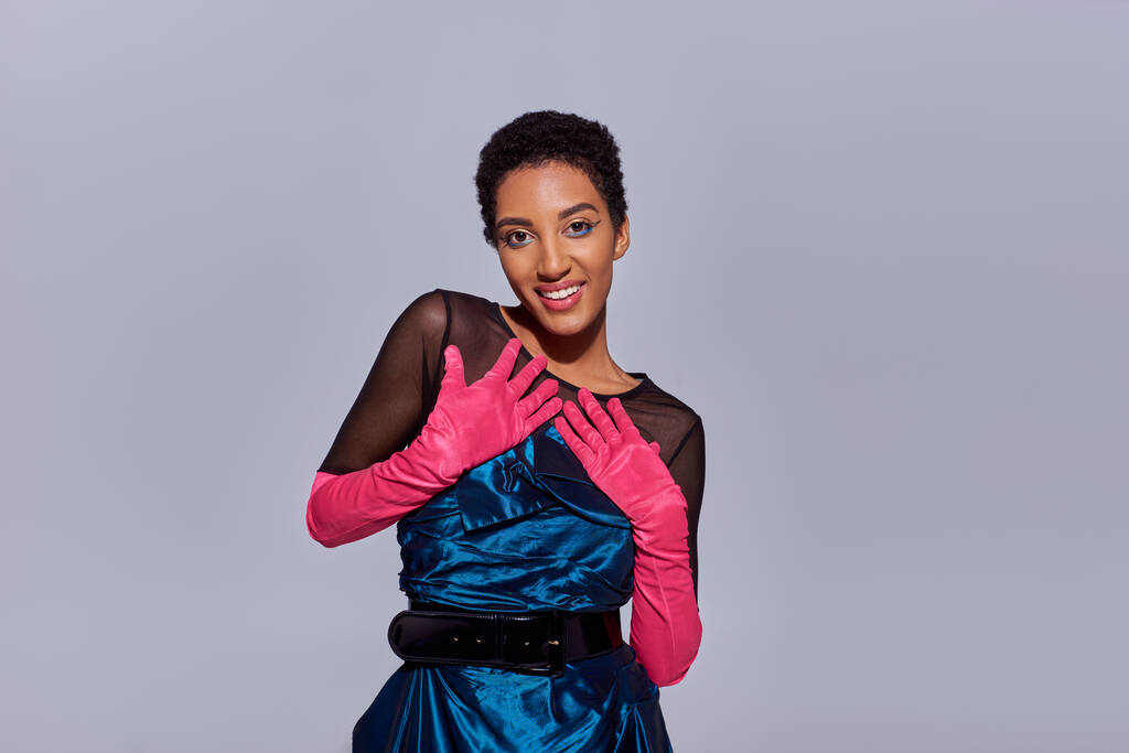 Portrét pozitivního a stylového afrického amerického modelu s výrazným make-upem v růžových rukavicích a koktejlových šatech při pohledu na kameru izolovanou na šedé, moderní módní koncepci generace z - Fotografie, Obrázek