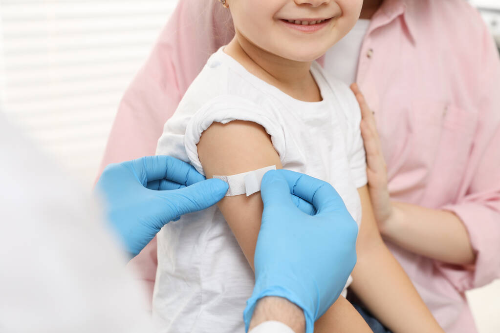 小児肝炎の予防接種。クリニックで娘と母。医者が女の子の腕に医療用の石膏を貼って - 写真・画像
