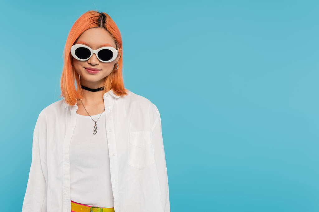 happy face, cheerful asian young woman with dyed hair standing in casual attire and sunglasses, smiling on blue background, white shirt, choker necklace, red hair, generation z  - Photo, Image