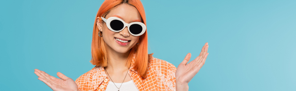positive, junge asiatische Frau mit gefärbten Haaren in lässiger Kleidung und Sonnenbrille, gestikulierend mit den Händen auf leuchtend blauem Hintergrund, orangefarbenem Hemd, Halskette, Generation z, rotem Haar, Banner - Foto, Bild