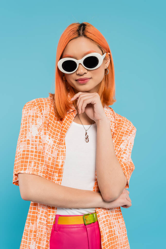 generación z, joven mujer asiática con el pelo teñido de pie en traje casual y gafas de sol, mirando a la cámara en el fondo azul vibrante, camisa naranja, pelo rojo, estilo personal - Foto, Imagen
