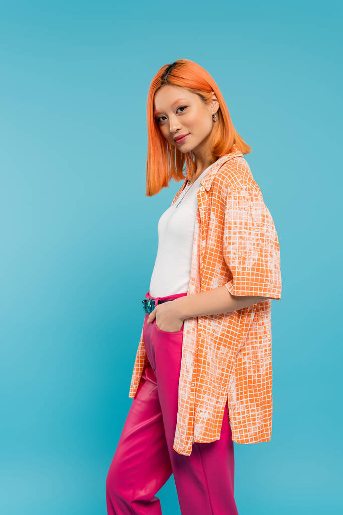 hand in pocket, pretty and young asian woman with dyed hair standing in orange shirt and posing on blue background, looking at camera, pink pants, generation z, modern style  - Photo, Image