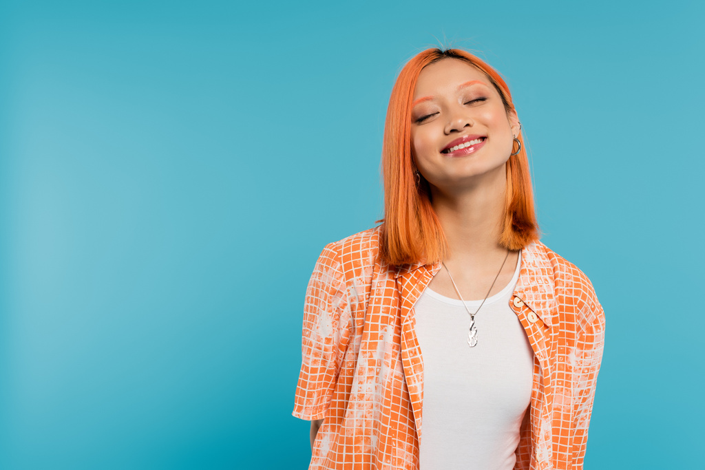 オレンジ色のシャツに閉じた目で髪の毛を染めた若いアジア人女性が青い背景で笑顔カジュアル服装幸せ自由陽気な態度  - 写真・画像
