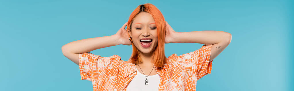 pozitivita a štěstí, mladá asijská žena s obarvenými vlasy stojící se zavřenýma očima v oranžové košili a usmívající se na modrém pozadí, ležérní oblečení, svoboda, veselý postoj, tetování, banner  - Fotografie, Obrázek