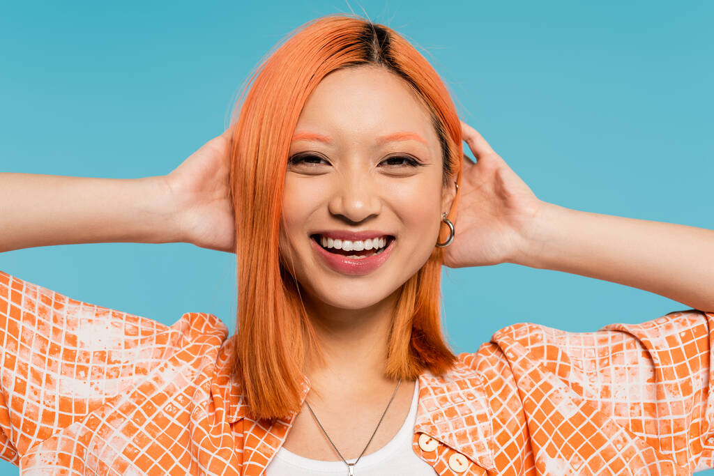 pozitivita, zářivý úsměv, mladá asijská žena s obarvenými vlasy stojící v oranžové košili a usmívající se na modrém pozadí, ležérní oblečení, svoboda, veselý postoj, při pohledu do kamery  - Fotografie, Obrázek