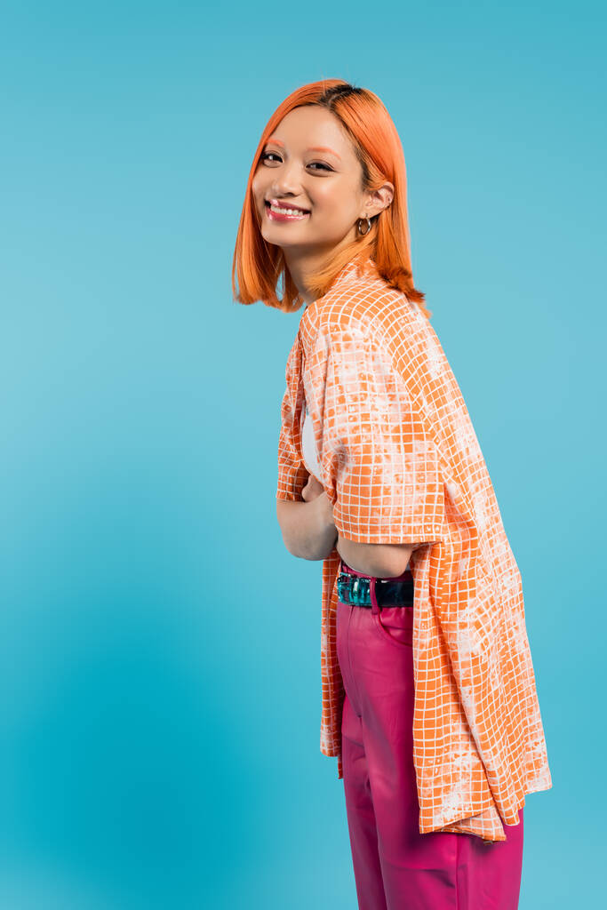 sorriso radioso, giovane donna asiatica con i capelli tinti in piedi con le braccia incrociate, in camicia arancione e sorridente su sfondo blu, abbigliamento casual, libertà, atteggiamento allegro, guardando la fotocamera, positività  - Foto, immagini